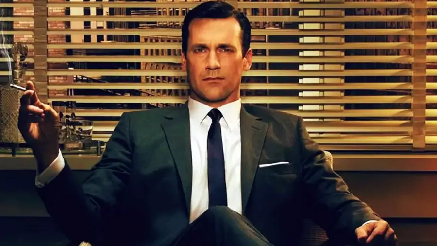 A man in a dark suit and tie sits confidently in an office chair, holding a cigarette. Behind him, Venetian blinds cover the window with city lights visible. The scene has a vintage, 1960s atmosphere.