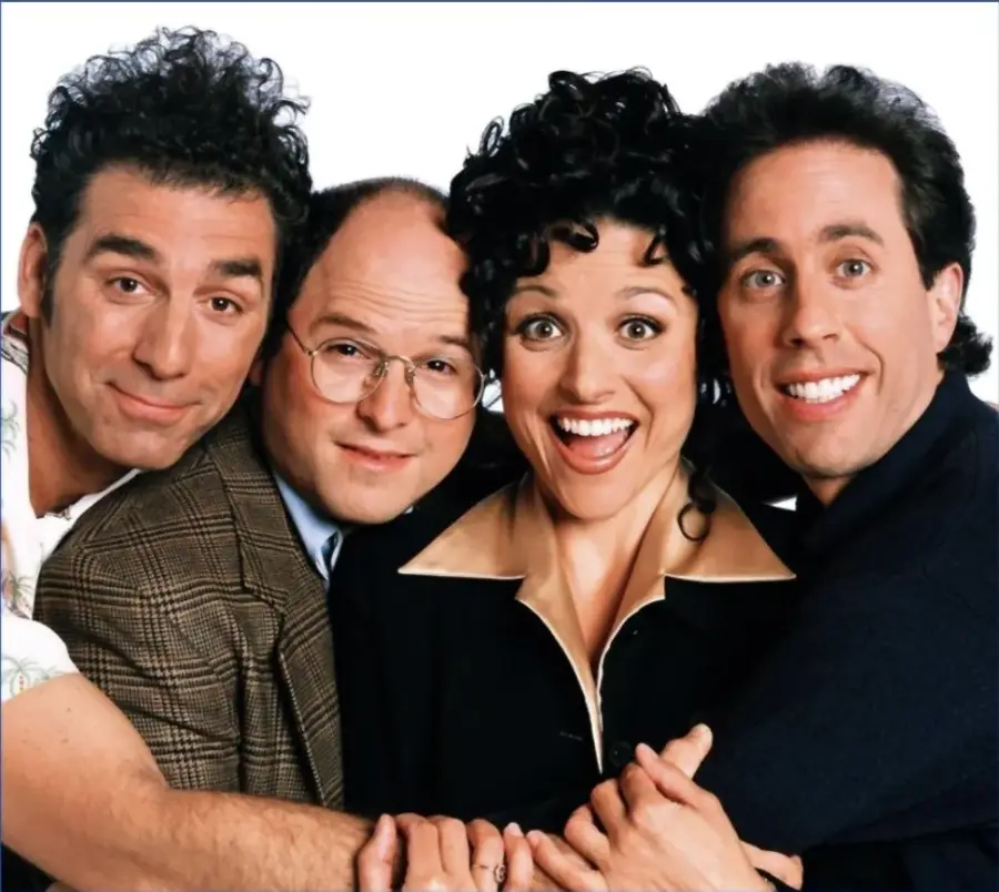Four people stand close together, smiling widely at the camera against a plain white background. They are casually dressed, with two men on the left, a woman in the center, and another man on the right.