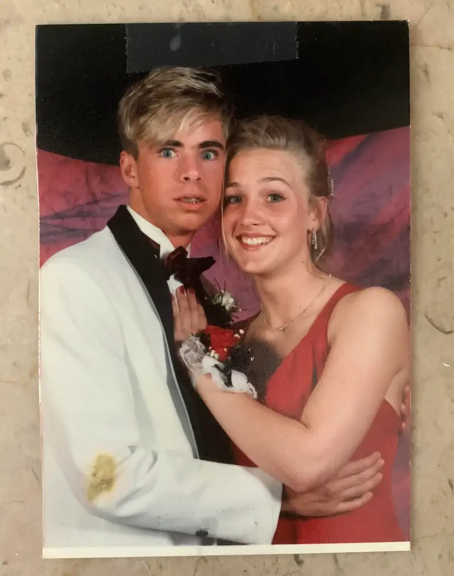 A young man in a white tuxedo jacket looks surprised while posing with a smiling young woman in a red dress at a formal event, possibly prom. They have corsages and stand in front of a purple backdrop.