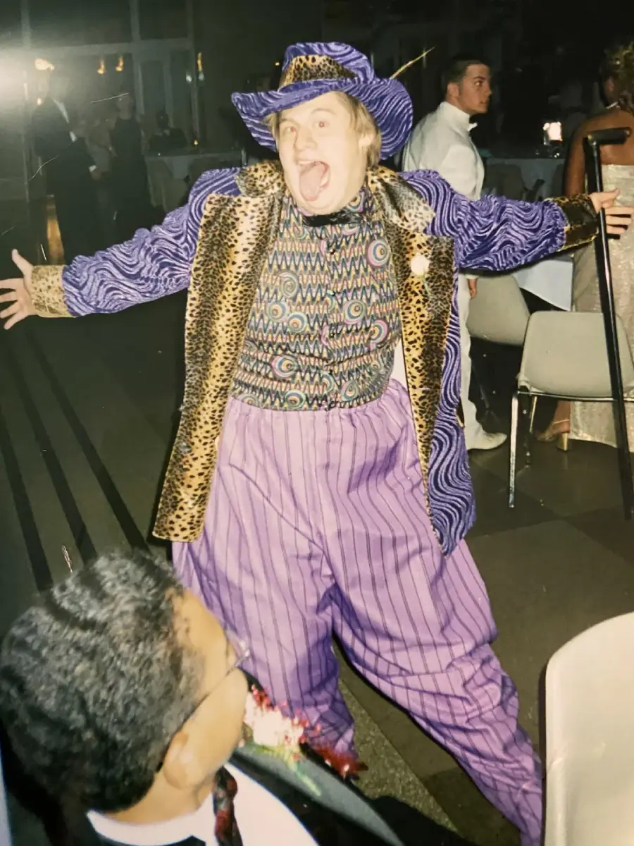 A person in a funky purple outfit with leopard print details and pinstripe pants poses energetically with arms wide open at an indoor event, while others sit at tables in the background.