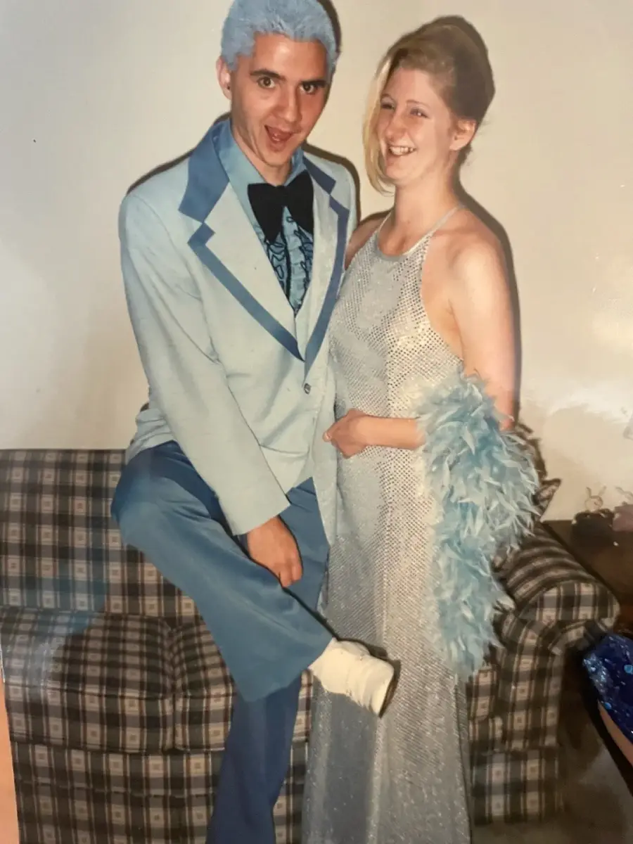 A man in a light blue tuxedo with matching dyed hair poses playfully next to a smiling woman in a silver dress with a blue feather boa, standing in front of a plaid couch indoors.