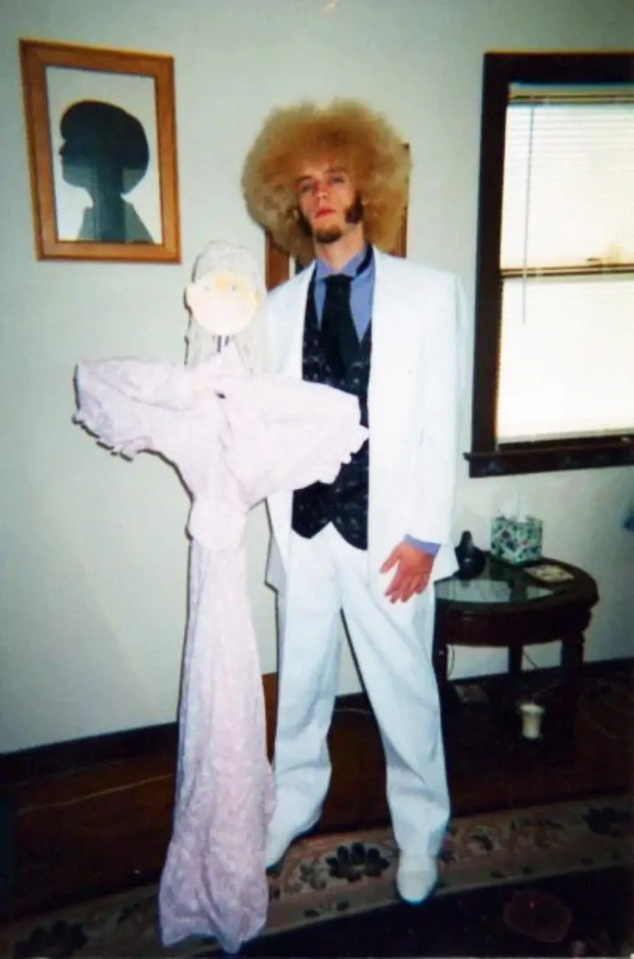 A man with a large blond afro wears a white suit and dark vest, posing indoors next to a figure made of pink fabric with a simple round face. A window and a framed silhouette picture are in the background.
