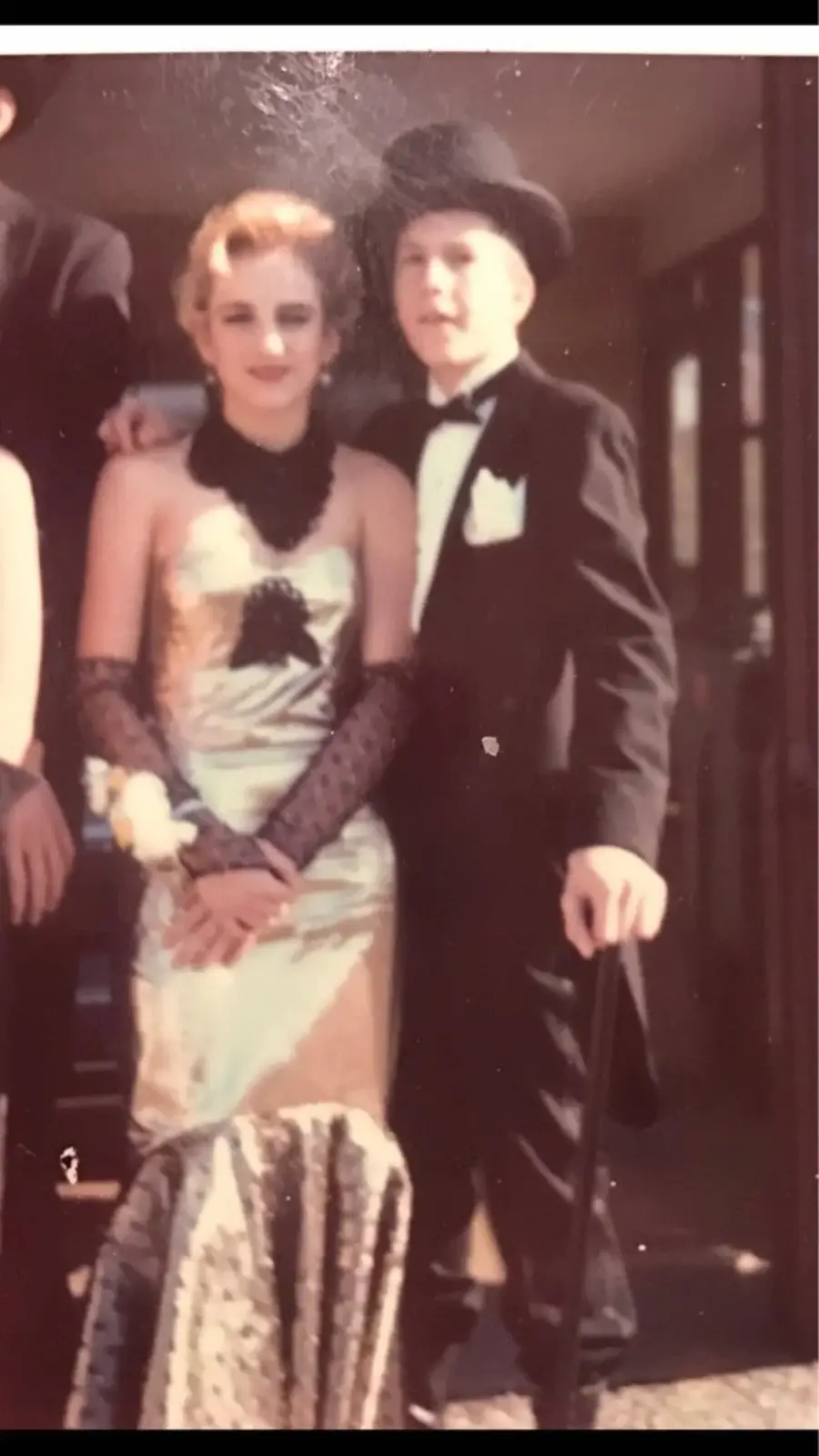 A young woman and man dressed in vintage formalwear pose together. The woman wears a strapless gown with black lace gloves, while the man wears a tuxedo with a top hat and cane. Both stand close, smiling for the photo.