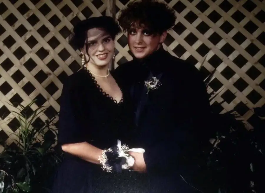 Two people dressed in formal dark clothing stand close together, smiling at the camera. They are in front of a wooden lattice backdrop with green plants. One person is wearing a veil and pearls, the other has a boutonnière.