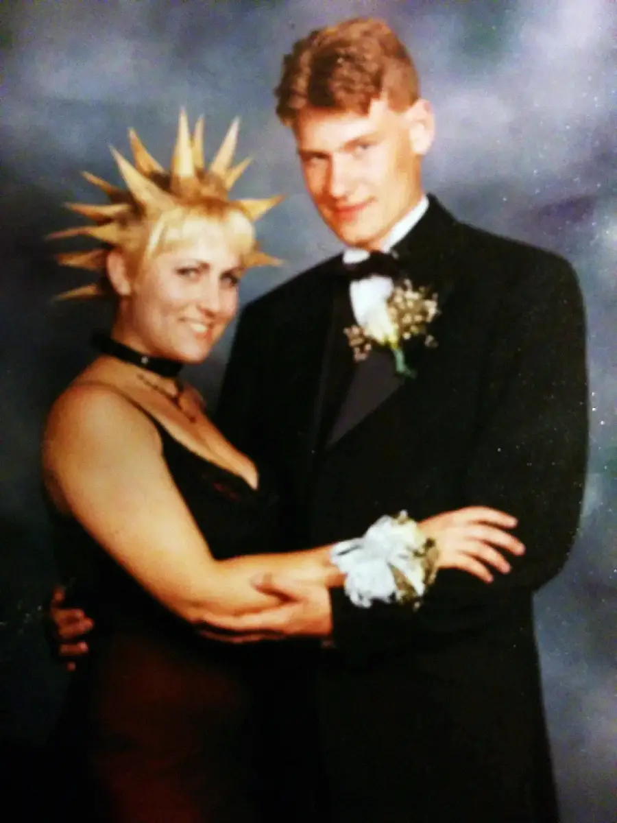 A couple in formal prom attire poses together; the woman has spiked blonde hair and wears a black dress with a choker, while the man wears a classic black tuxedo with a bow tie and boutonnière.