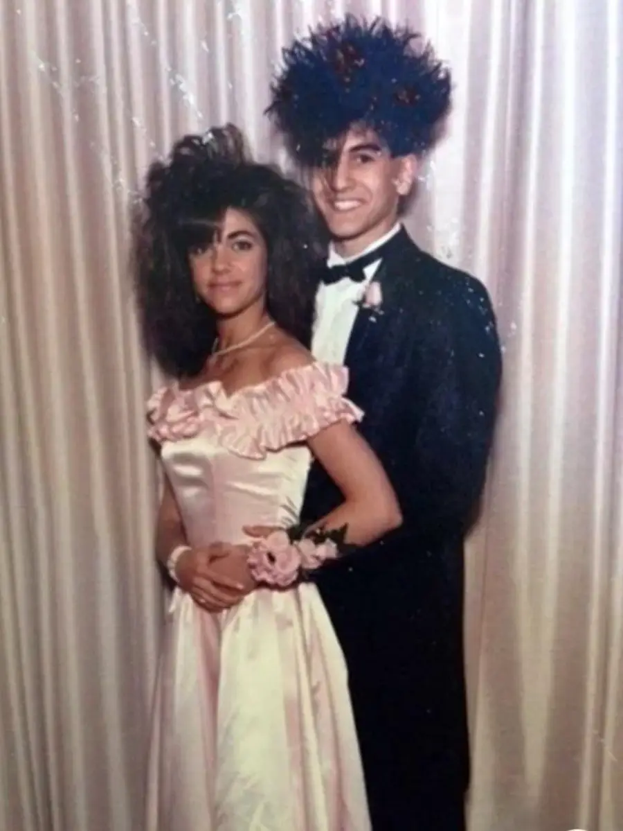 A young couple poses for a formal photo in front of a curtain; the woman wears a pink off-shoulder dress with a corsage, and both have big, teased 1980s-style hair. The man is in a black tuxedo and bow tie.