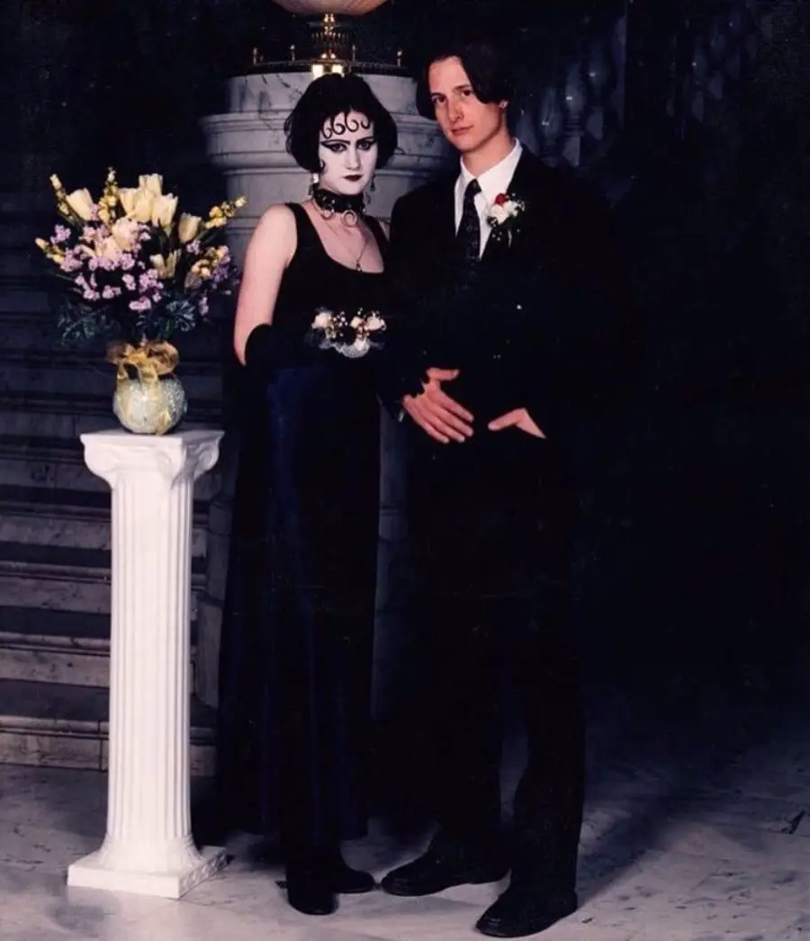 Two people in formal attire pose together; the person on the left wears a dark gothic dress with dramatic makeup and jewelry, while the person on the right wears a black suit. They stand next to a white pedestal with a bouquet of flowers.