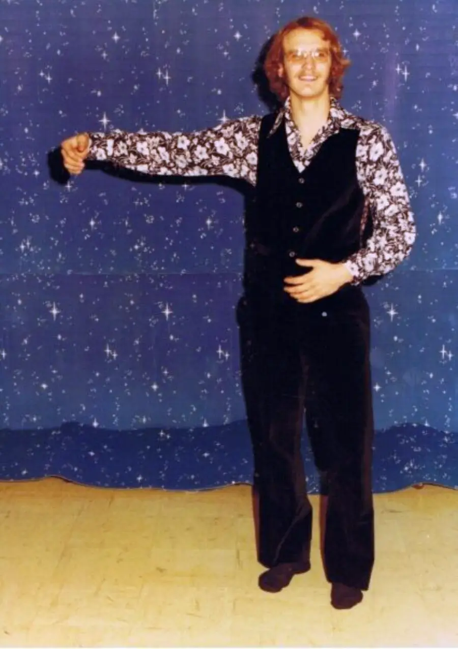 A smiling person wearing glasses, a black vest, and floral-patterned shirt stands on a wooden floor against a blue starry backdrop, posing with one arm bent and the other outstretched as if holding an invisible partner.