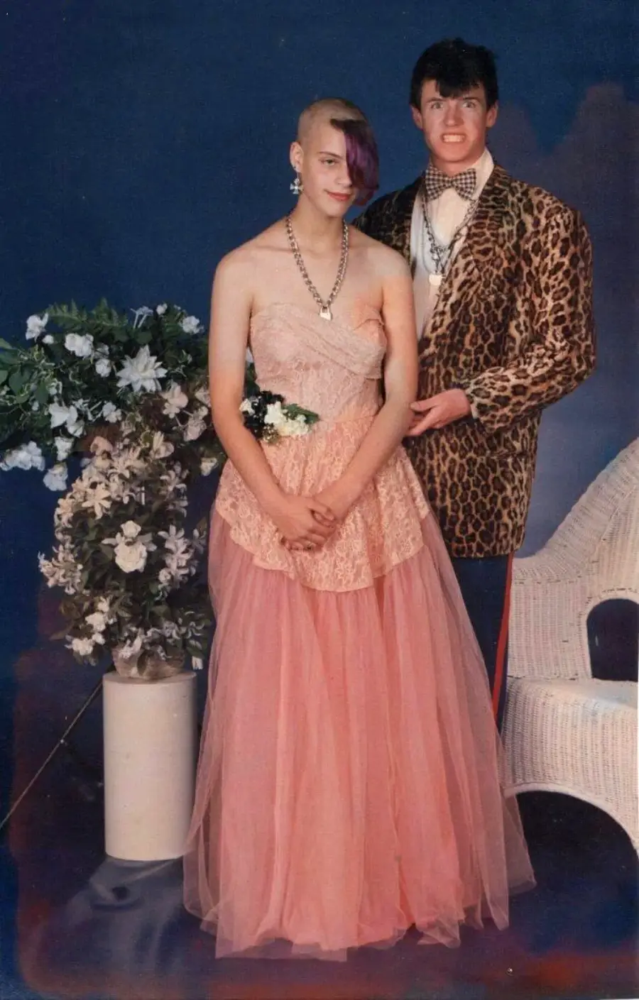 A young couple poses for a formal photo; the woman wears a pink lace gown with a floral corsage, and the man wears a leopard print jacket, bow tie, and makes a funny face. They stand by a wicker chair and a vase of white flowers.