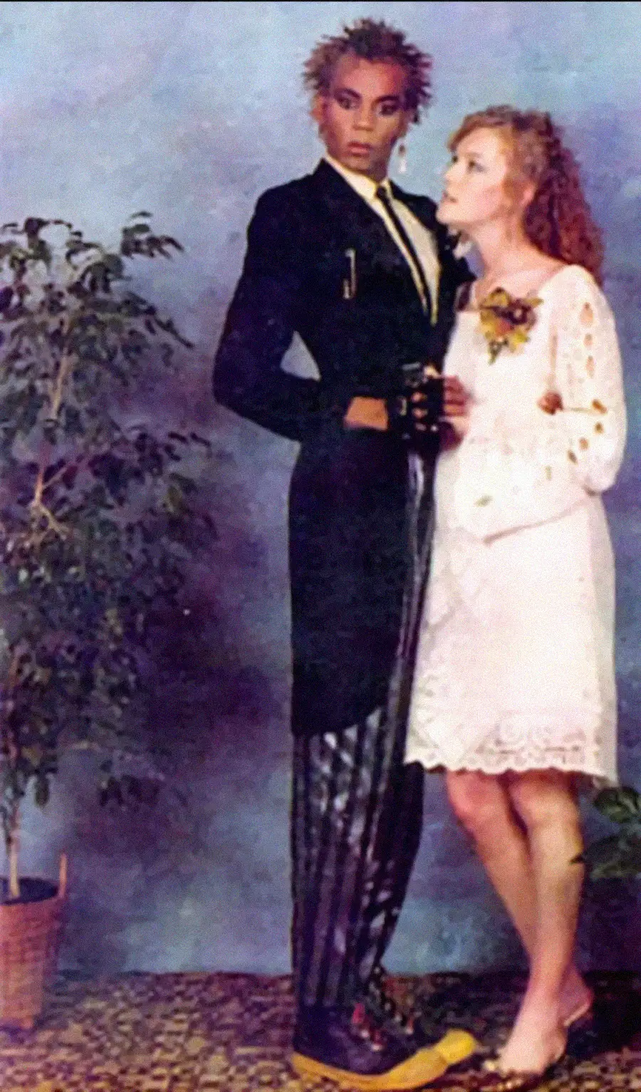 A young couple poses for a formal photo. The person on the left wears a black jacket, striped pants, and yellow shoes, with spiked hair. The person on the right wears a white lace dress and holds a corsage, standing close together.