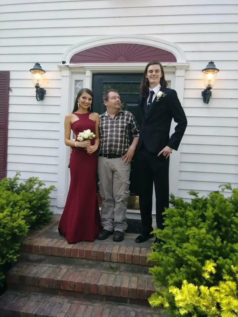 Three people stand on brick steps in front of a white house. A woman in a red dress holds flowers, a man in a plaid shirt stands in the middle, and a tall man in a black suit stands to the right. Shrubs border the steps.
