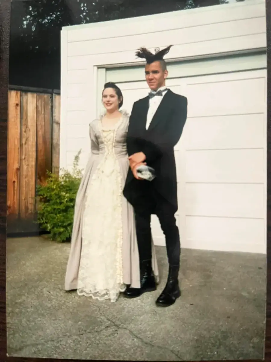 A young couple poses outside in formal attire; the woman wears a cream and gray vintage-style gown, while the man wears a black suit with a dramatic, tall hairstyle that fans upward. They stand on pavement in front of a garage door.