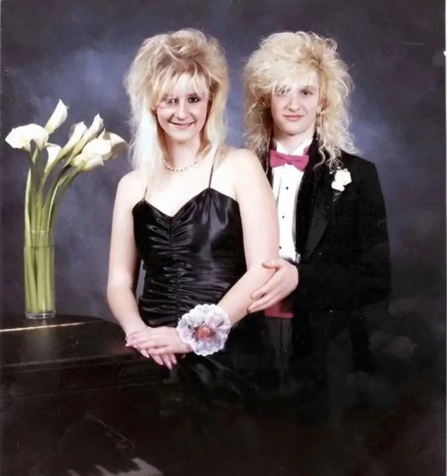 Two people with big, teased blonde hair pose for a formal photo. One wears a black dress with a corsage; the other wears a tuxedo with a pink bow tie. They stand next to a vase of white calla lilies against a dark backdrop.