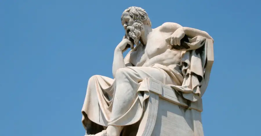 A marble statue of a seated, bearded man with a contemplative expression, resting his chin on one hand, against a clear blue sky.
