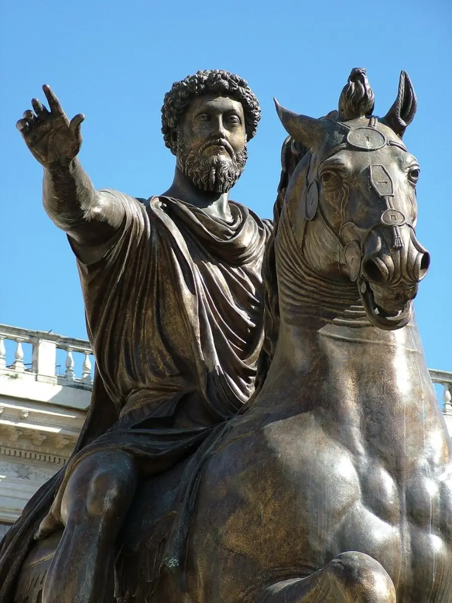 A bronze statue of a bearded man with curly hair in a toga, seated on a horse with one arm raised. The statue stands outdoors with a blue sky and part of a building in the background.
