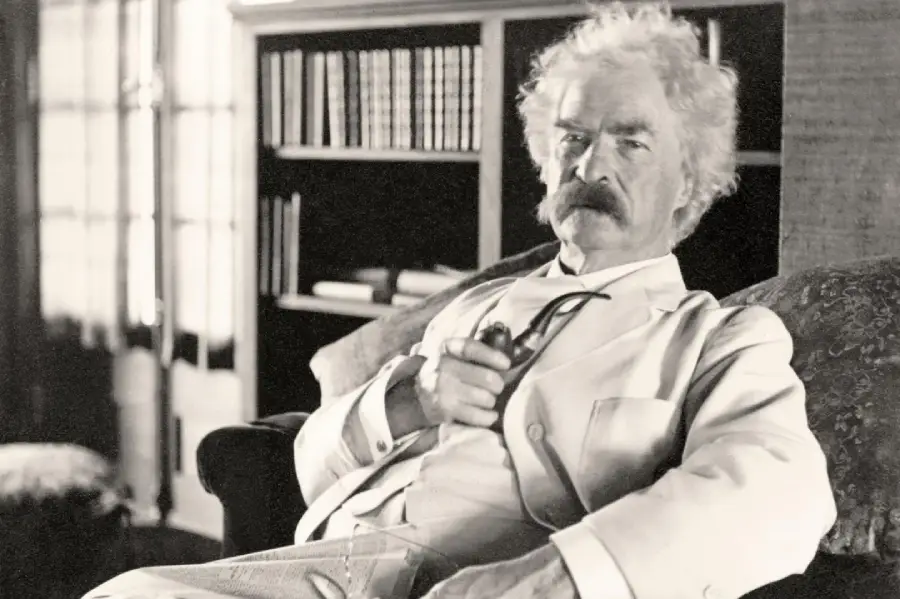 An older man with white hair and a bushy mustache sits in an armchair, wearing a light-colored suit and holding a pipe. A bookcase filled with books is in the background.