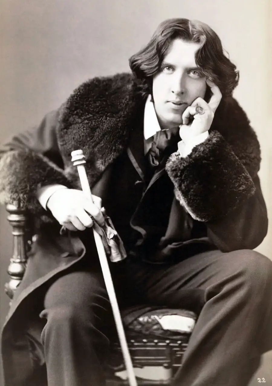 A man in formal Victorian-era clothing sits on a chair, holding a cane and resting his head on his hand, gazing thoughtfully. He has wavy, dark hair and wears a fur-collared coat.