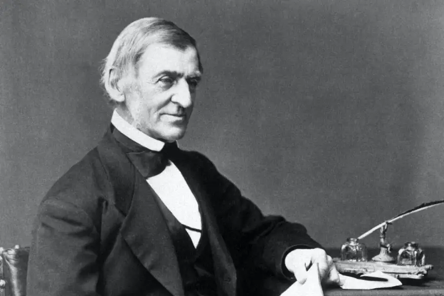 Black-and-white photo of an older man in 19th-century formal attire, seated at a desk with papers, an inkwell, and a quill pen, looking slightly to his left with a calm expression.