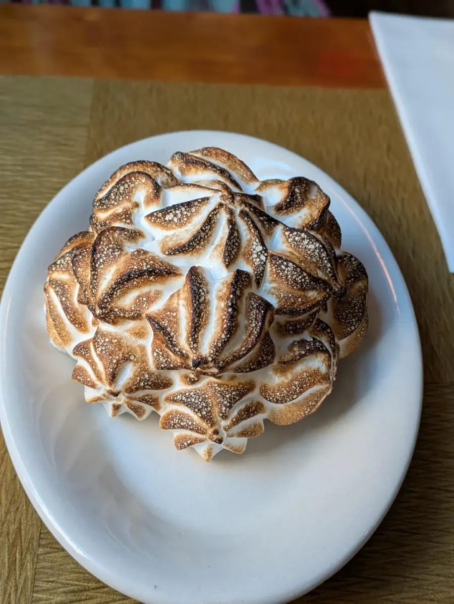 A dessert topped with intricately piped and toasted meringue, shaped into flower-like patterns, served on a white plate on a wooden table.