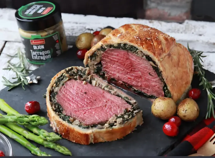 A sliced Beef Wellington with a golden pastry crust, pink beef center, and a layer of mushrooms and greens, garnished with asparagus, roasted potatoes, cherry tomatoes, and a jar of Dijon tarragon mustard.