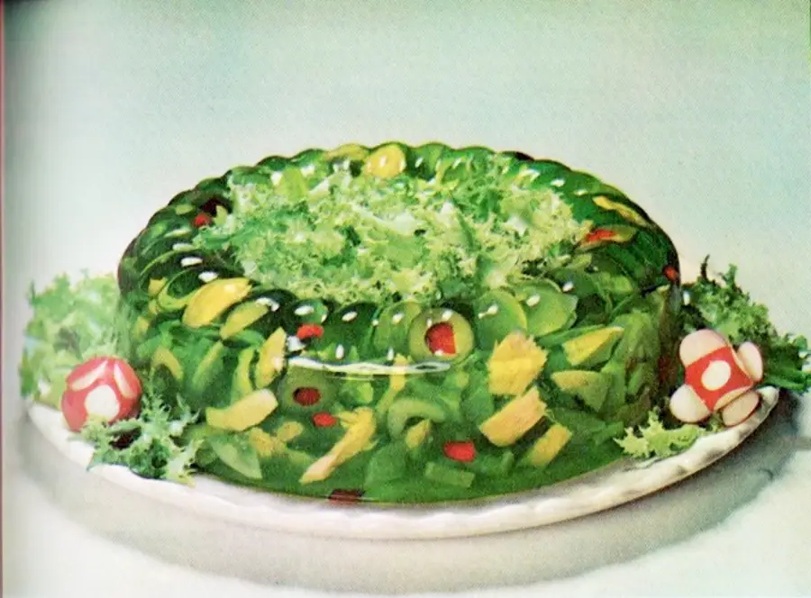 A retro-style green gelatin mold filled with vegetables sits on a plate, garnished with leafy greens and two radish-and-vegetable decorations resembling small mushrooms.