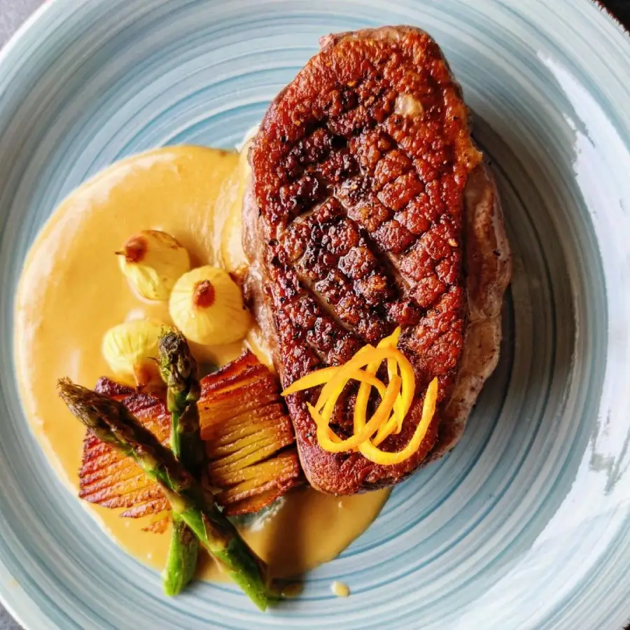 A gourmet dish with seared duck breast, golden brown and scored on top, served with creamy sauce, roasted potatoes, glazed pearl onions, asparagus spears, and a garnish of citrus zest, presented on a blue ceramic plate.