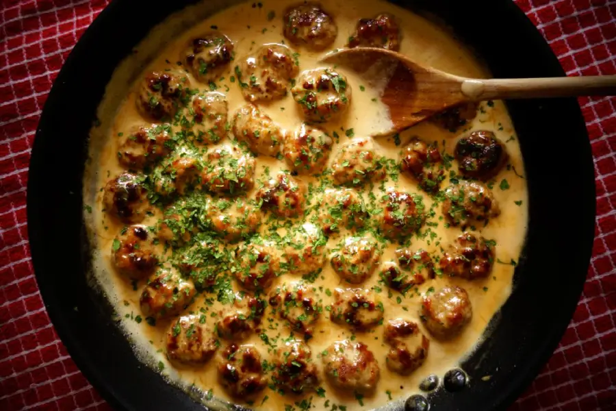 A skillet filled with meatballs in a creamy sauce, garnished with chopped herbs, sits on a red checkered cloth. A wooden spoon rests on the edge of the skillet.