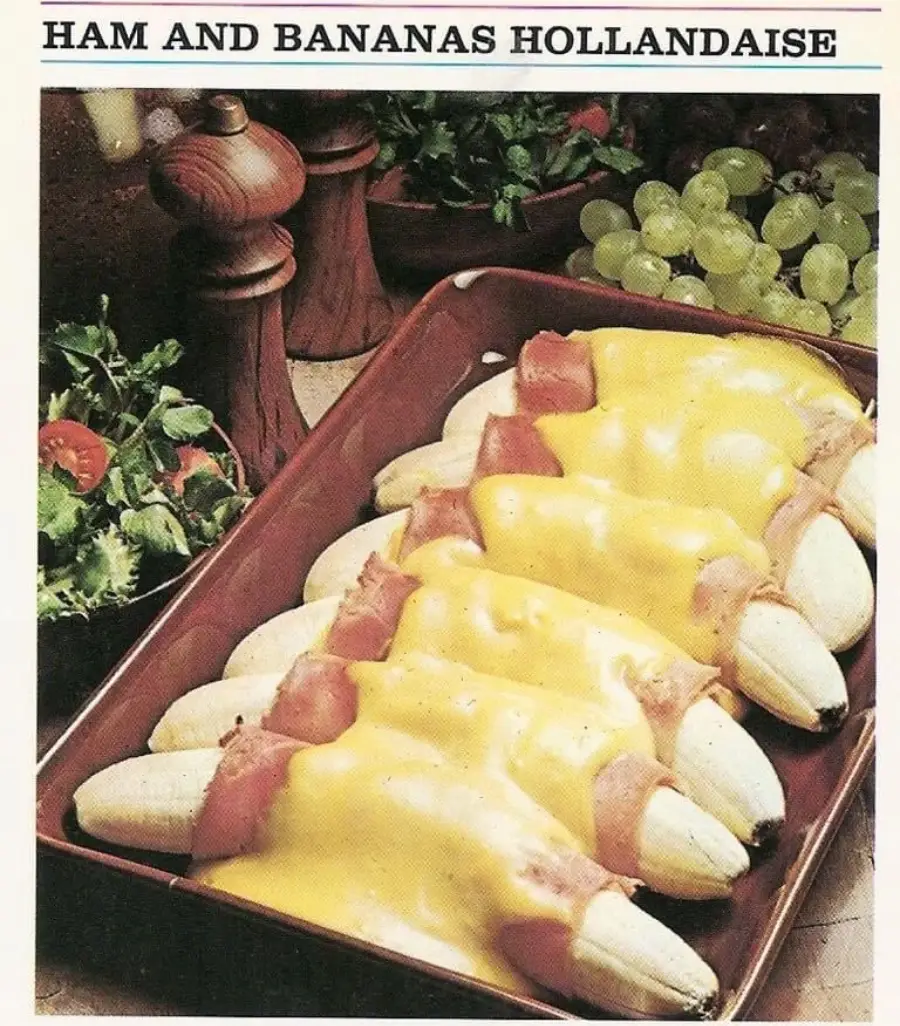 A dish of peeled bananas wrapped in ham, topped with yellow hollandaise sauce, is arranged in a brown baking dish. Grapes, parsley, and pepper mills are visible in the background. The title reads "Ham and Bananas Hollandaise.