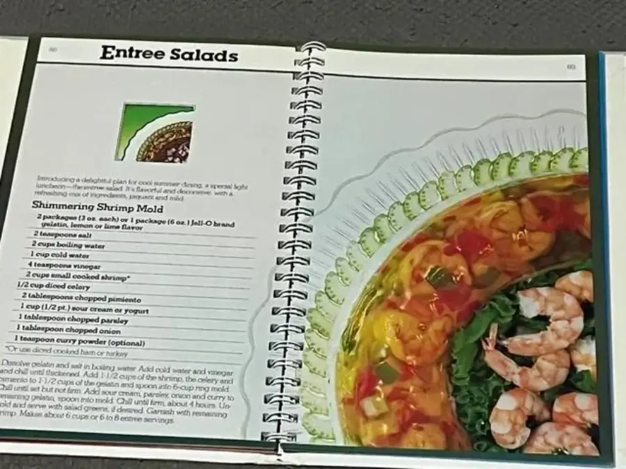 An open spiral-bound cookbook shows a recipe for "Shimmering Shrimp Mold" on the left and a photo of a colorful shrimp gelatin mold on a clear platter on the right. The shrimp are arranged atop greens.