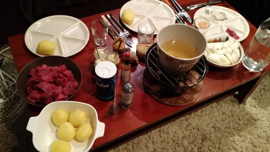 A table set for a meal with divided plates, forks, and glasses, featuring raw beef cubes, peeled potatoes, sliced onions, condiments, and a pot of broth on a burner, ready for fondue.