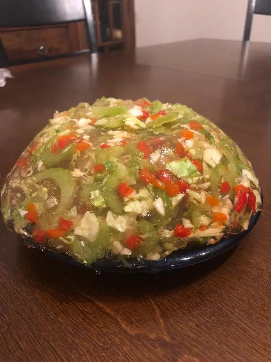 A dome-shaped gelatin salad filled with chopped celery, red bell peppers, and other vegetables sits on a blue plate on a wooden table.