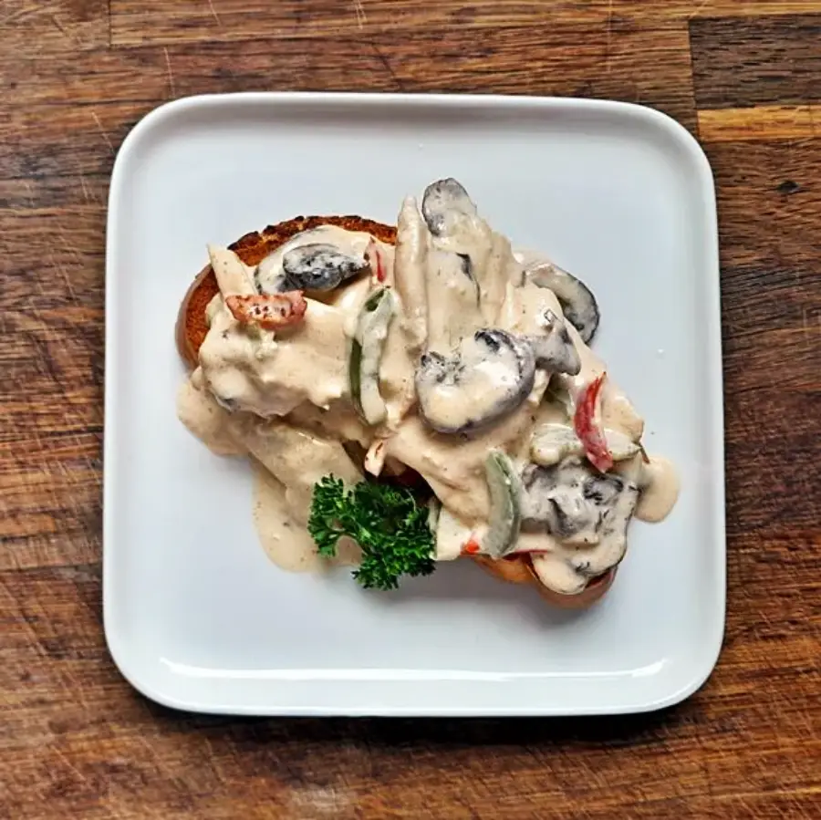A slice of toast topped with creamy mushroom and vegetable sauce, garnished with a sprig of parsley, served on a white square plate over a wooden surface.