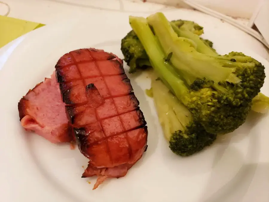 A white plate with two slices of grilled ham, marked with grill lines, next to a serving of steamed broccoli.