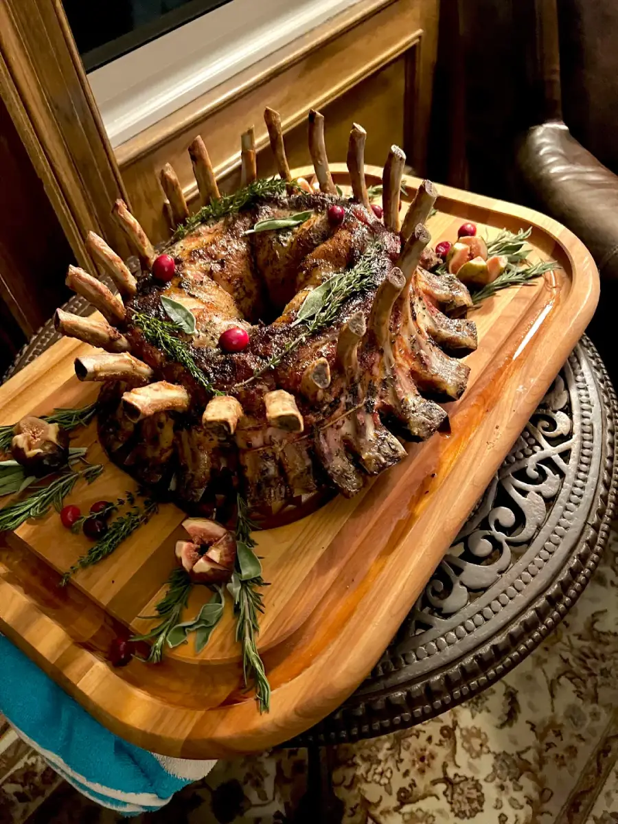 A beautifully roasted crown rack of pork garnished with fresh herbs and cranberries, arranged on a wooden serving board with sprigs of rosemary and halved figs. The board rests on an ornate round table.