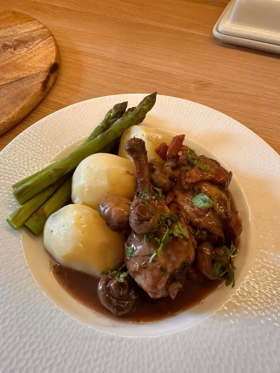 A plate of coq au vin with mushrooms and bacon, served with boiled potatoes and steamed asparagus spears, on a white textured plate on a wooden table.