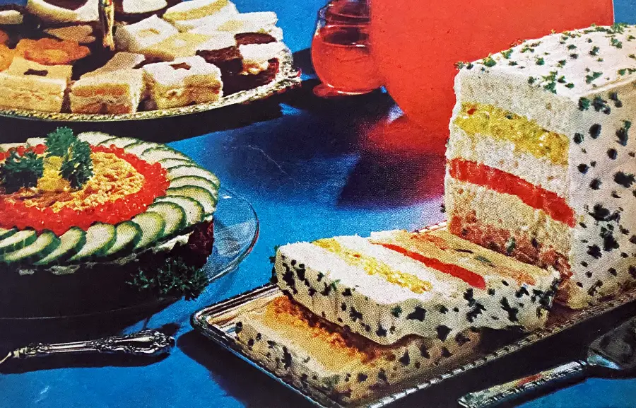 A retro-style spread of party foods on a blue table, featuring a layered savory loaf covered in herbs, a round dish topped with sliced cucumber and tomato, and assorted square sandwiches on a tray in the background.