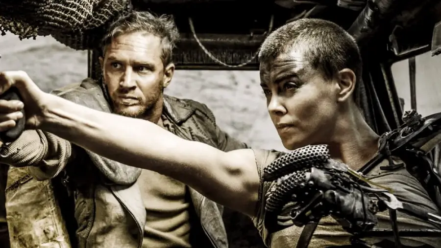 Two people with intense expressions sit inside a rugged vehicle; the woman has a shaved head and is aiming a gun, while the man looks alert beside her. They are both dressed in worn, post-apocalyptic clothing.