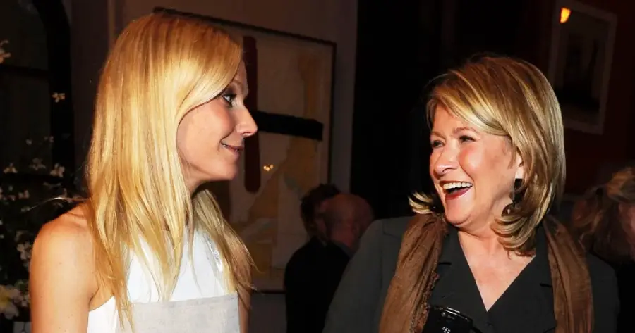 Two women standing together at an indoor event, smiling and engaged in conversation. One has long blonde hair and wears a white top; the other has shoulder-length blonde hair and wears a dark outfit with a scarf.