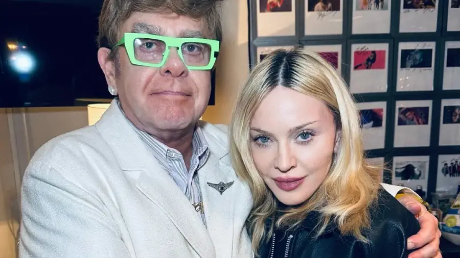 Two people pose together indoors. The person on the left wears light green glasses, a white jacket, and a collared shirt. The person on the right has long blonde hair and a black jacket. Framed photos are visible on the wall behind them.