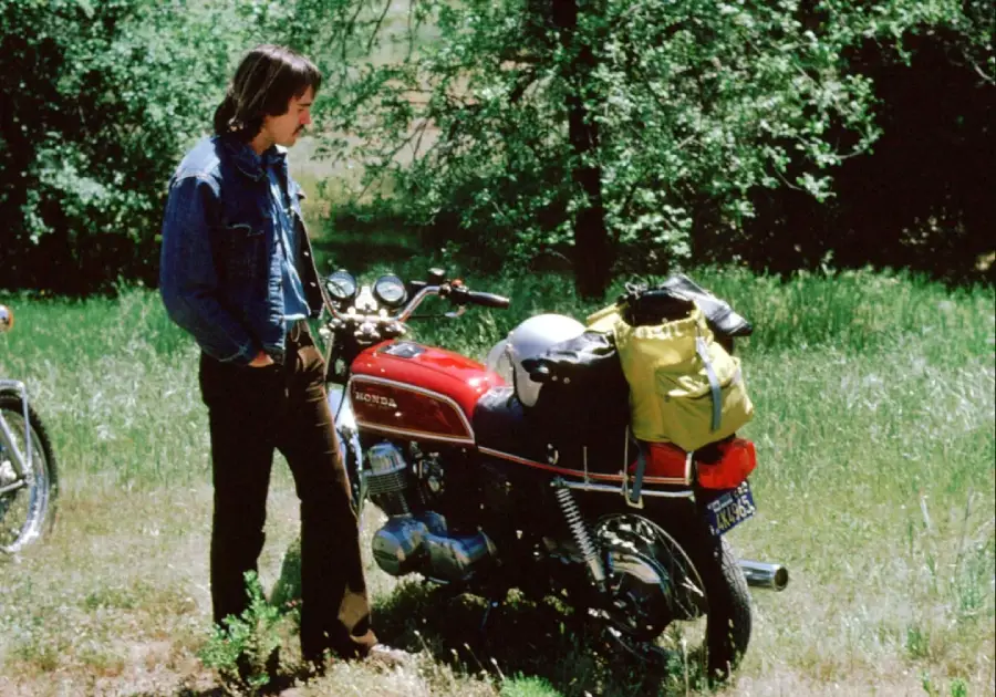 A person in a denim jacket stands next to a red Honda motorcycle with a yellow bag strapped to the back, parked on grass with green trees in the background.