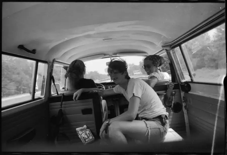 Three young people ride in a vintage car. One sits in the back, looking at the camera, while two face forward in the front seats. A carton of milk rests beside them. Trees and road are visible through the windows.