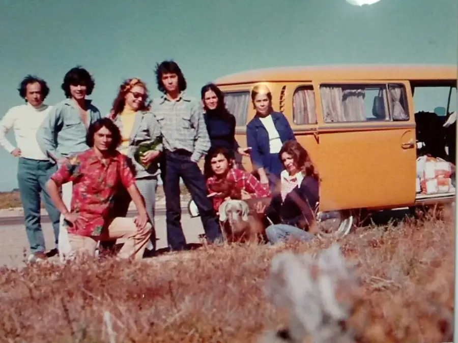 A group of eight people pose outdoors by an orange van on a sunny day. Some are standing and some are kneeling in front, with bags visible inside the van. Dry grass and open sky are in the background.
