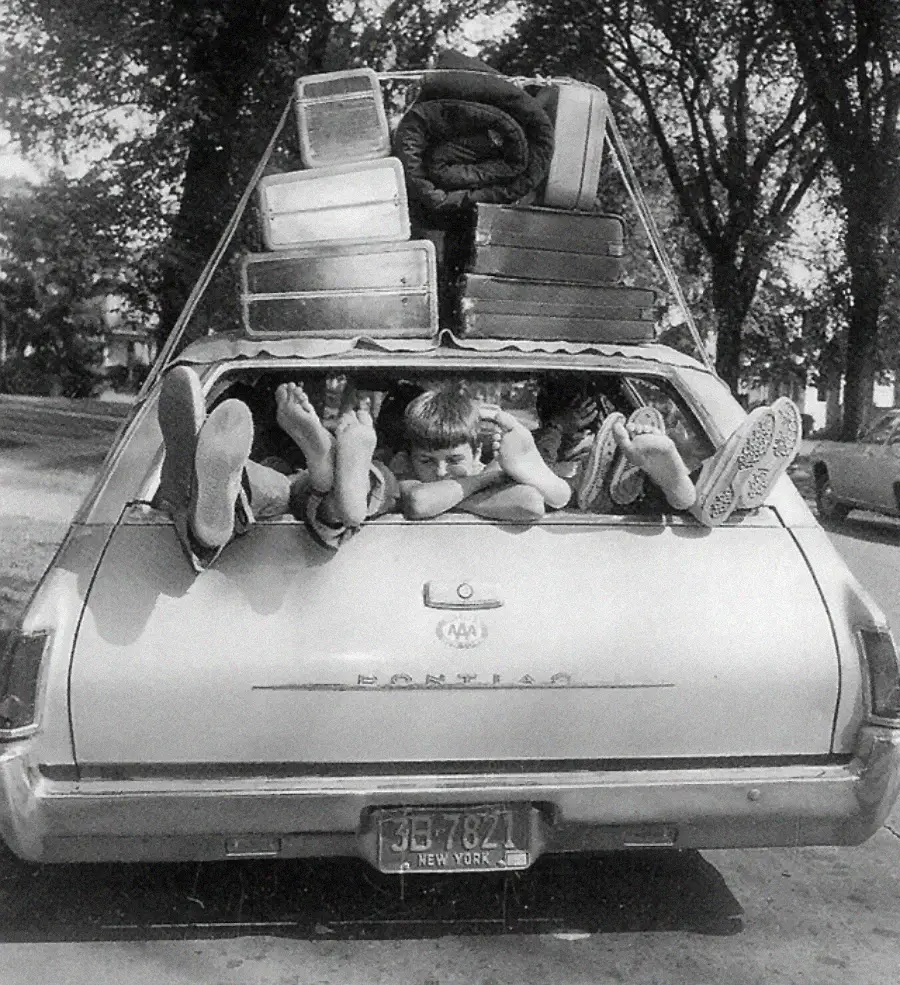 Three children are lying in the open back of a station wagon with their feet sticking out, surrounded by luggage stacked on the car’s roof. The scene appears playful, with trees visible in the background.