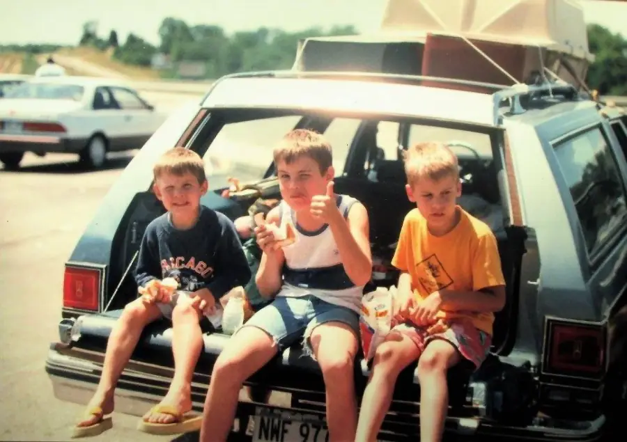 Three young boys sit on the open tailgate of a station wagon, eating snacks. The car is parked in a lot with another car and trees in the background. The boys appear to be on a road trip, with luggage on the car roof.