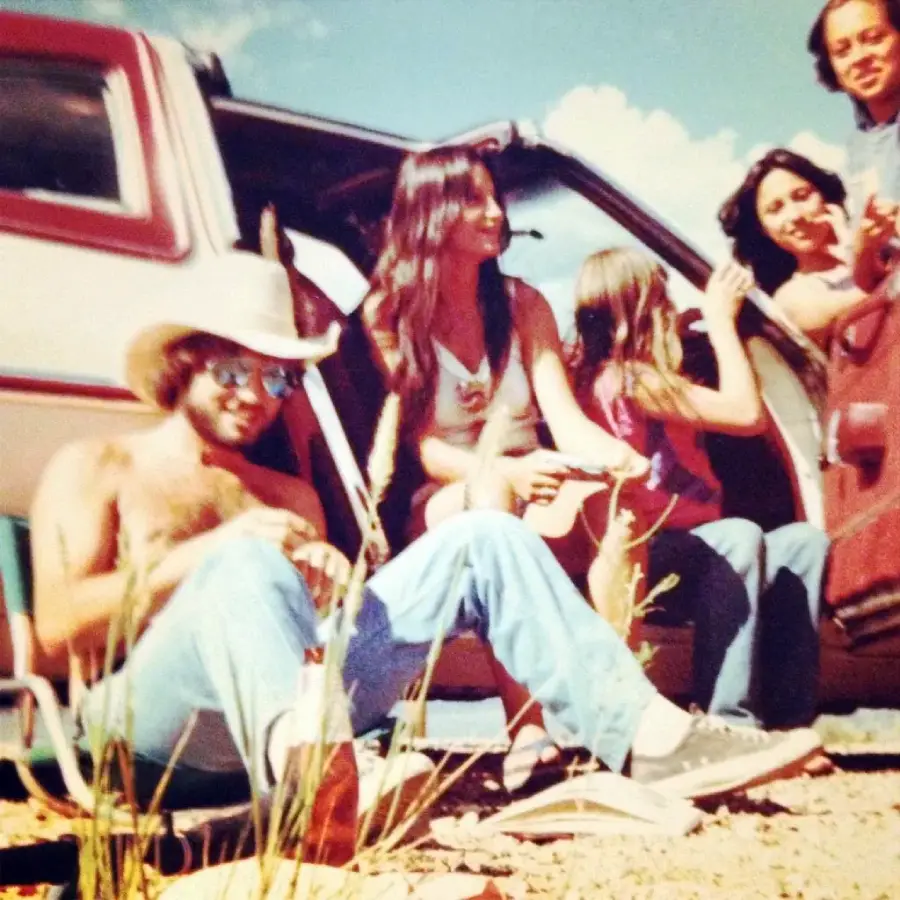 A group of people relax by an open car door outdoors on a sunny day. One man wears a cowboy hat and sunglasses, while others sit or kneel nearby, smiling and talking. Tall grass and rocks are in the foreground.