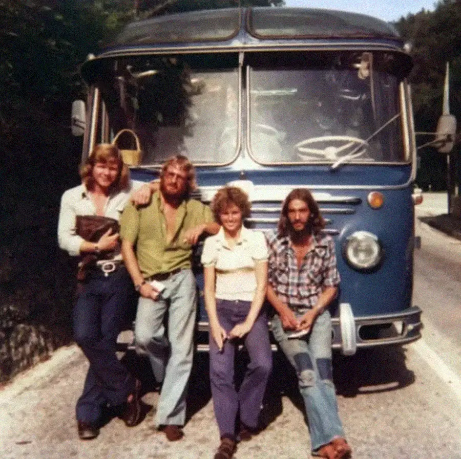Four people are sitting and leaning against the front of a vintage blue bus parked on a road, posing for the photo. The group appears relaxed and is dressed in casual 1970s-style clothing.