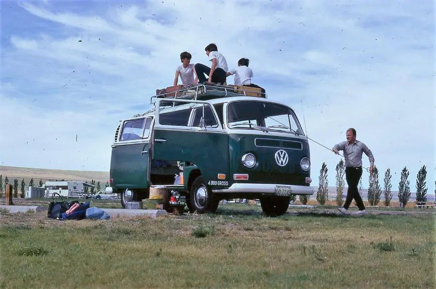 A vintage green Volkswagen van is parked on grass with its side door open. Three people sit on the roof, while another person stands nearby holding onto a rope tied to the van. Camping gear is scattered on the ground.