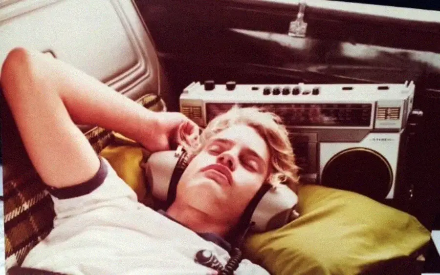 A young man with blond hair is lying down with large headphones on, eyes closed, and resting his head on a green pillow. A vintage boombox is visible next to him.