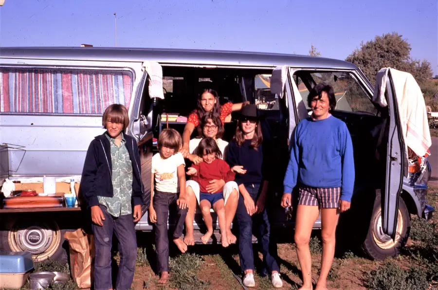A group of seven children and teens pose smiling in front of a blue van with curtains. Some sit in the open doorway, while others stand barefoot on grass, with camping items visible around the van on a sunny day.