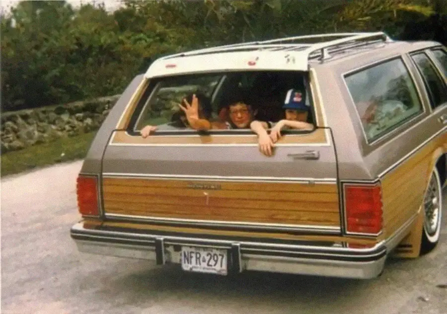 Three kids look out the open rear window of a vintage station wagon with wood paneling. One waves at the camera. The car is parked outdoors with trees and a stone wall in the background.